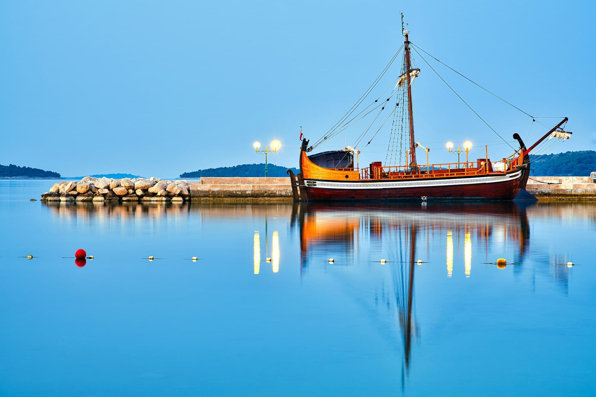 Bonaca, a traditional wooden boat with a tall mast and an orange hull, mirrored in a flat calm harbour