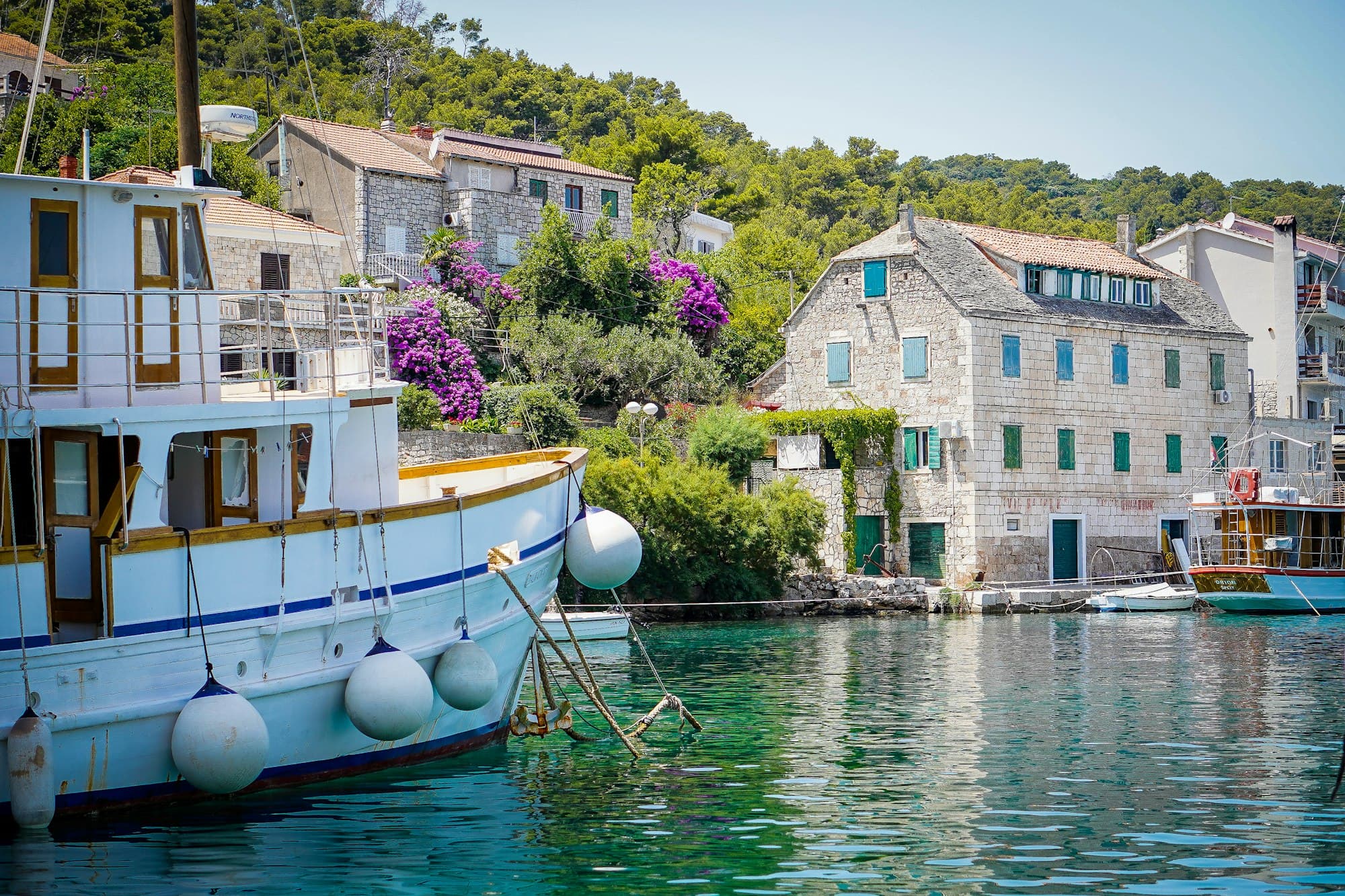 Galeb, a small white wooden boat with a blue stripe, tied up below stone houses and bougainvillea